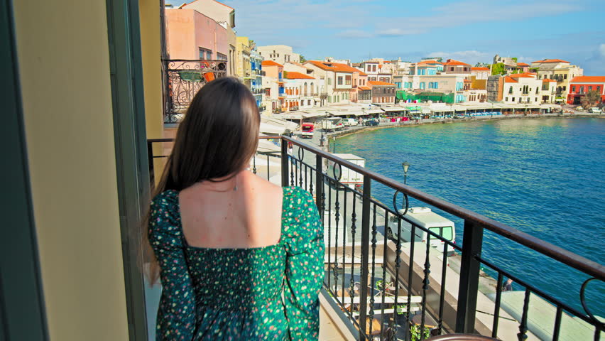 A beautiful girl in a dress enjoying the view from the hotel balcony in Chania, Crete. Young woman overlooking colourful Old Venetian Port of Chania surrounded by turquoise-blue water, mosque landmark