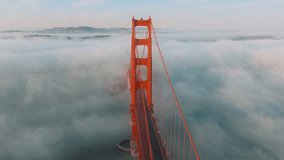Karl the Fog drifting below red bridge main landmark of San Francisco slow motion California USA. Aerial video cinematic Golden Gate Bridge. Cars driving by red bridge covered in scenic white clouds