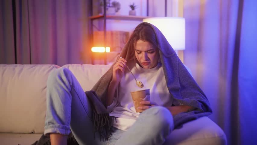 Portrait of sad young woman dealing with stress by eating food sitting on couch at home Upset brunette female wrapped in blanket crying and eating ice cream indoors at late night Mental heath problem Royalty-Free Stock Footage #1110732761
