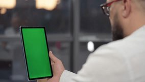 Bearded Man in Glasses Using Tablet Computer with Green Screen Mock Up Display. Male Watching Videos and Reading Social Media Posts on Mobile Device. Shooting Close Up Over the Shoulder