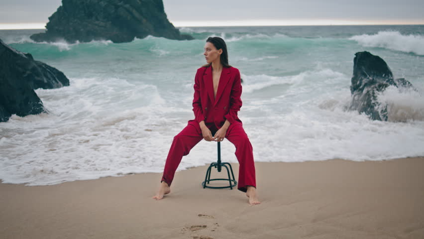 Stylish beautiful lady sitting on beach washed bare legs by foamy waves. Attractive confident model posing at chair near rock gray dramatic ocean view. Sensual young woman wearing red elegant suit.