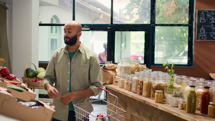 Middle eastern man in eco food store examining organic locally grown fruits and veggies, opening jars with additives free pasta or homemade sauces. Client admires fresh bio merchandise.