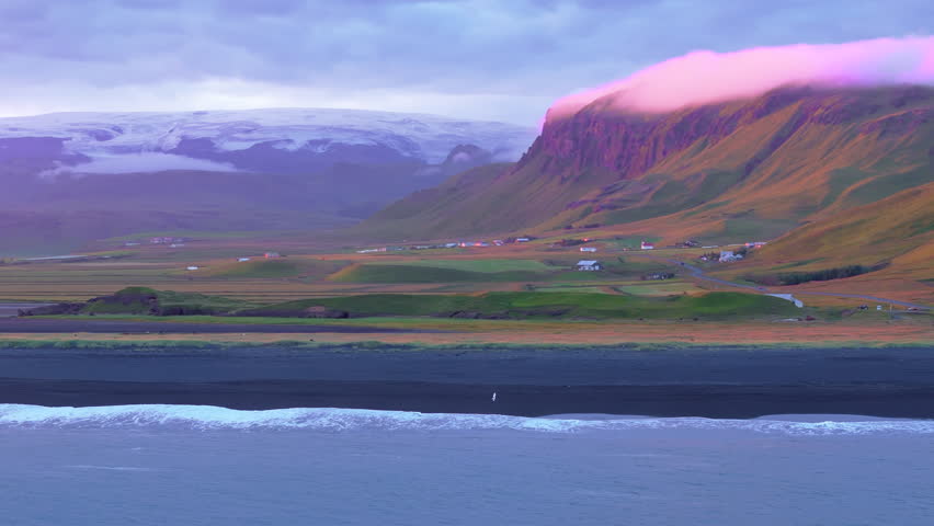 Ariel shot of running man in white on Reynisfjara Black Sand Beach in Iceland during unbelievable sunset with purple cloudy sky and snowy mountains on the background and green Hills on the foreground Royalty-Free Stock Footage #1110754439