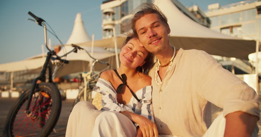 Portrait of a couple: A girl in light clothes leans on the shoulder of a guy with stubble in a white beach shirt against the background of white cafe umbrellas. A guy and a girl rode a bicycle to the