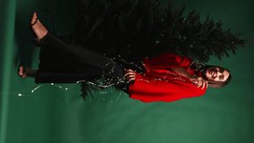 a girl in a red sweater has garlands wrapped around herself and is posing against a Christmas tree and a green background. vertical video