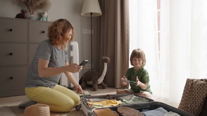 Mom and son work as team, packing their suitcase for upcoming trip. With excitement and determination, they carefully fold clothes and check off items on their list. Advertising trip mobile app Royalty-Free Stock Footage #1110760929