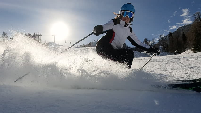 LENS FLARE, SLOW MOTION Recreational skier sprays snow while skiing on ski track. Experienced female skier is carving on slopes of ski resort in snowy Austrian Alps. Nice sunny day for winter sports.