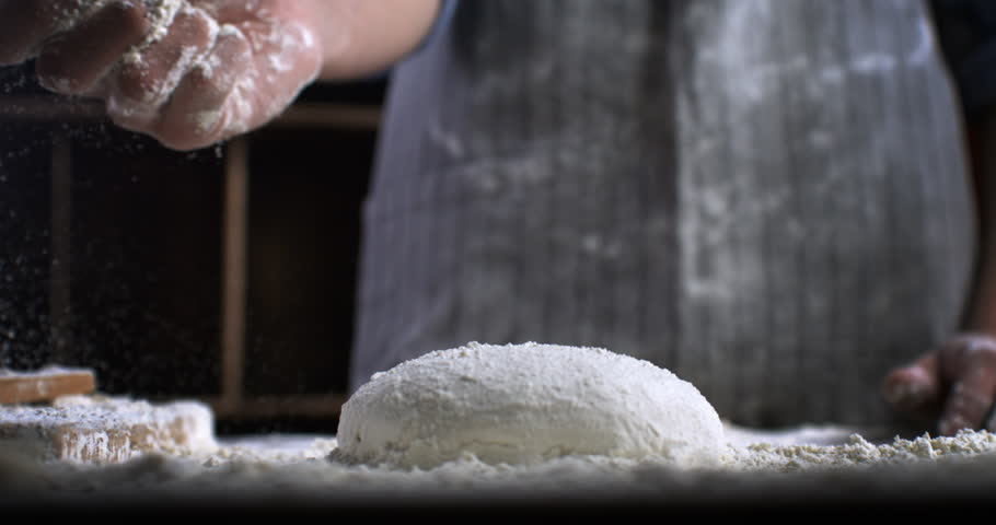 Super slow motion close up of professional artisan baker chef sprinkles flour on raw loaf of dough while making homemade bread, pasta or pizza on rustic wooden table in traditional bakery kitchen. Royalty-Free Stock Footage #1110762859