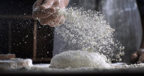 Super slow motion close up of professional artisan baker chef sprinkles flour on raw loaf of dough while making homemade bread, pasta or pizza on rustic wooden table in traditional bakery kitchen. Stockvideó