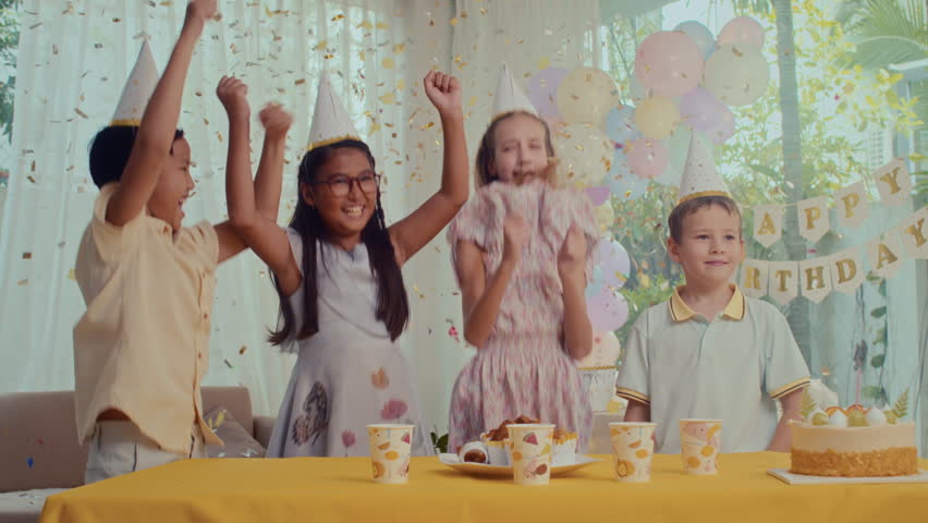 Medium long shot of happy diverse kids in party hats jumping in confetti next to festive table while celebrating birthday at home Royalty-Free Stock Footage #1110763757