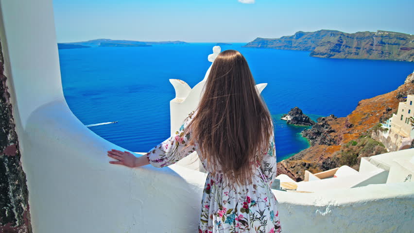 Beautiful girl visiting Santorini and enjoying the view of blue roofs, hotels and resorts. Happy woman walking on the streets of the famous blue dome church in the village of Cyclades island, Greece.