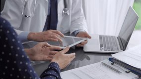 Doctor and patient are sitting at the grey glass table in clinic. Male pediatrician is using a tablet computer while discussing something with a middle-aged woman. Medicine concept