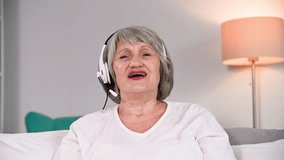 portrait of a cute old pensioner talking on a video call using a headset while sitting in a cozy room, looking at camera