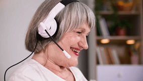 portrait of a smiling elderly woman using headphones talking online via a web camera on a laptop while relaxing at home, smiling and looking at the camera