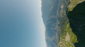 Vertical video. FPV sport drone dynamic dive shot close to natural rock sunlight valley green grass high altitude. Summer light ski resort mountain summit with cable car suburb aerial panorama view 4k