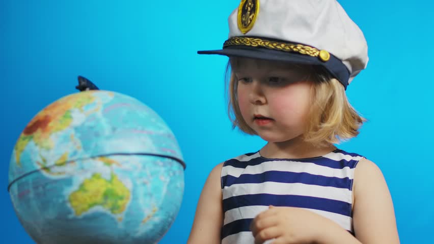 Little cute girl in a sailor hat is pointing her finger into the globe and smiles on the blue background in the studio