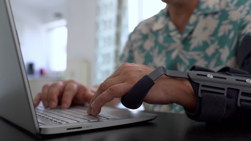 Man with elbow injury, special modern adjustable articulated elbow orthosis typing on laptop keyboard. Side view shot man wearing arm splint sitting at desk in front window working distantly at home