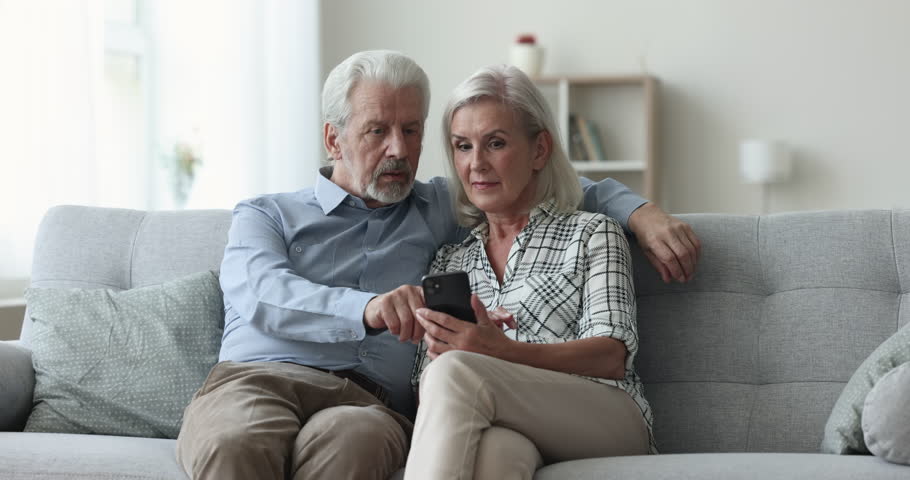 Focused elder retired husband and wife browsing messages on mobile phone, getting excited, happy, screaming for joy with winner hands, celebrating victory, triumph, win, enjoying entertainment Royalty-Free Stock Footage #1110791369