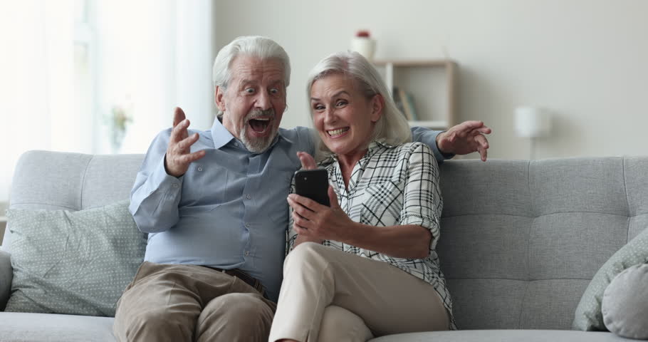 Focused elder retired husband and wife browsing messages on mobile phone, getting excited, happy, screaming for joy with winner hands, celebrating victory, triumph, win, enjoying entertainment Royalty-Free Stock Footage #1110791369