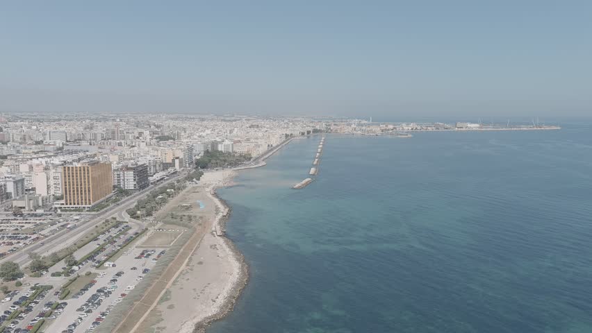 Bari, Italy. The Lungomare di Bari seafront promenade stretches along the Adriatic coast, captured in a sweeping aerial view that reveals the full scale of this historic waterfront. The camera glides over calm turquoise waters toward a dense urban skyline of white and pale limestone buildings that line the shore. A long breakwater extends into the sea, creating a protected harbor area, while a sandy beach and coastal road run parallel to the water