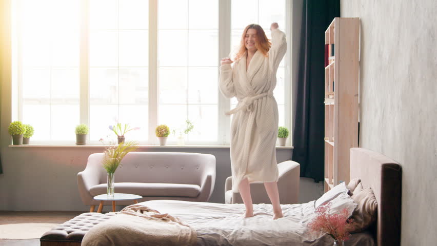 Home morning party in bedroom cheerful woman having fun carefree Caucasian happy active girl in bathrobe dancing jumping on bed moving listen to music dance jump joy move holiday in luxury hotel room Royalty-Free Stock Footage #1110797575