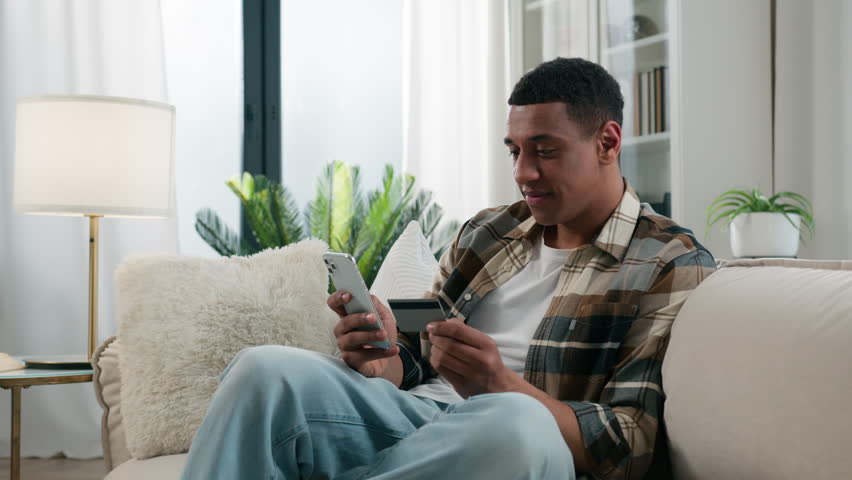 Happy African American man at home couch sofa guy user buyer male client banking buying shopping online with credit bank card and mobile phone smartphone e-commerce order delivery payment internet pay Royalty-Free Stock Footage #1110797609