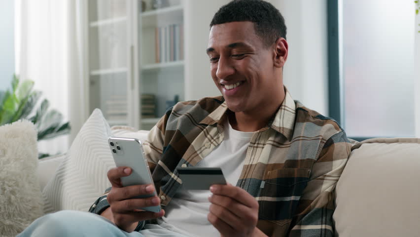 Happy African American man at home couch sofa guy user buyer male client banking buying shopping online with credit bank card and mobile phone smartphone e-commerce order delivery payment internet pay Royalty-Free Stock Footage #1110797609