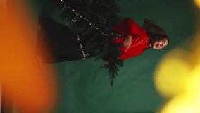 in the foreground bokeh garlands. a girl in a red sweater holds a Christmas tree in her hands and plays it like a guitar. the girl is wrapped in garlands. vertical video