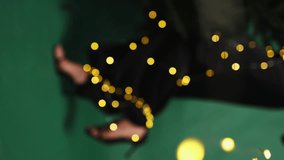 female legs in heels and a black dress on a green background wrapped in glowing yellow garlands. bokeh garland. vertical video