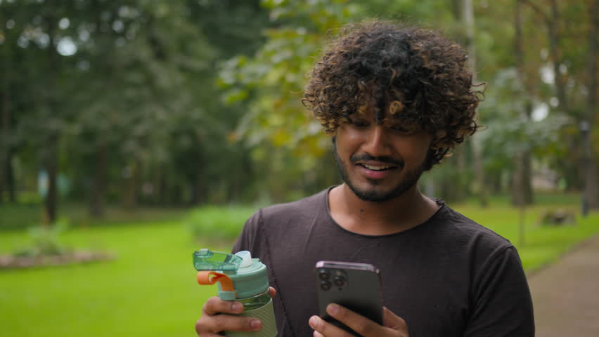 Fitness man Arabian guy walking in city park hold sport bottle water relax after training healthy workout outside exercise break happy smiling Indian sportsman using mobile phone walk with smartphone Royalty-Free Stock Footage #1110811197