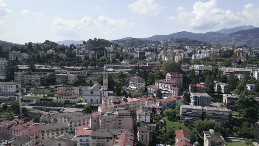 Aerial dolly over streets and buildings of Lugano, Switzerland. City center, red rooftops, moutains in the background, train station.