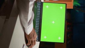 Vertical video Arabic man scrolling on mockup laptop while working from home. Self employed person writing emails on isolated screen device in apartment office with TV as background noise