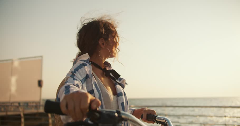 A happy brown-haired girl walks along the beach along the sea and carries her bicycle next to her. Girl on a bike ride near the sea Royalty-Free Stock Footage #1110826329