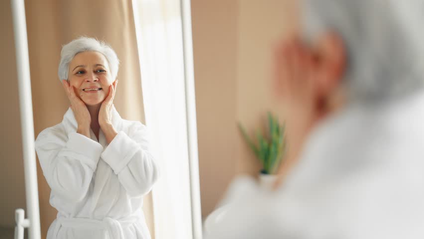 Senior wellbeing smiling woman in bathrobe with gray hair looking at mirror touching face worrying about aging process. Good looking old lady searching face wrinkles. Getting old age change concept. - Powered by Shutterstock - Get 15% off with code: PIKWIZARD15