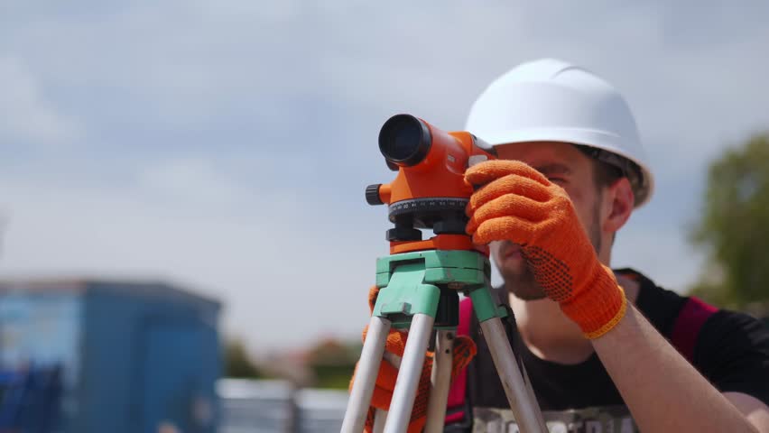 Construction worker uses optical level on construction site. Civil engineer with a theodolite doing land surveying work. Man wearing hard hat and work gloves measures angles for building plans. Royalty-Free Stock Footage #1110858545