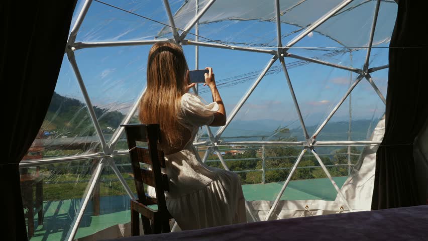 Young woman captures mountain view using smartphone inside geodesic dome accommodation, scenic nature backdrop.