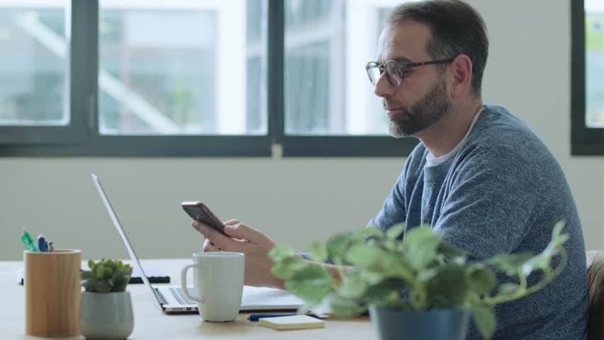 Businessman sitting at desk with laptop looking at smartphone - Powered by Shutterstock - Get 15% off with code: PIKWIZARD15
