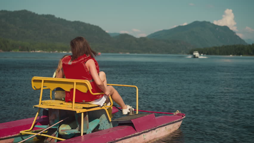 Young woman in red life jacket sits in catamaran looking at boat. Tourist enjoys landscape on clear lake slow motion backside view. Summer fun on water in recreation area