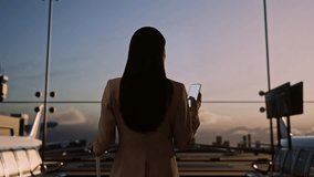 Back View Of Asian Businesswoman With Rolling Suitcase In Boarding Lounge At The Airport, Using Smartphone, Walking And Looking Around, Airplane Takes Off Outside The Window - Powered by Shutterstock - Get 15% off with code: PIKWIZARD15