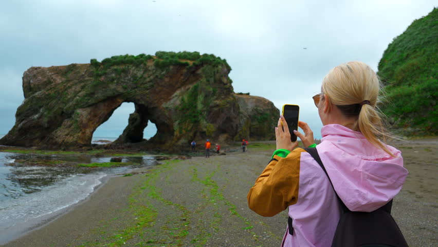 Woman on shore takes pictures of sea on phone. Clip. Woman photographs seashore with rocky arch. Group of tourists takes pictures of rocky arch on seashore