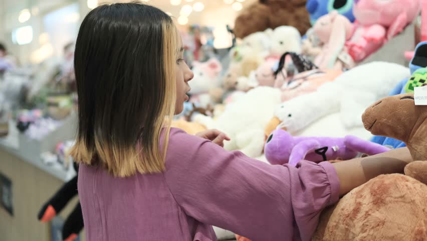 A little girl in a lilac elegant dress against the background of soft toys in a store. A child chooses a toy as a gift