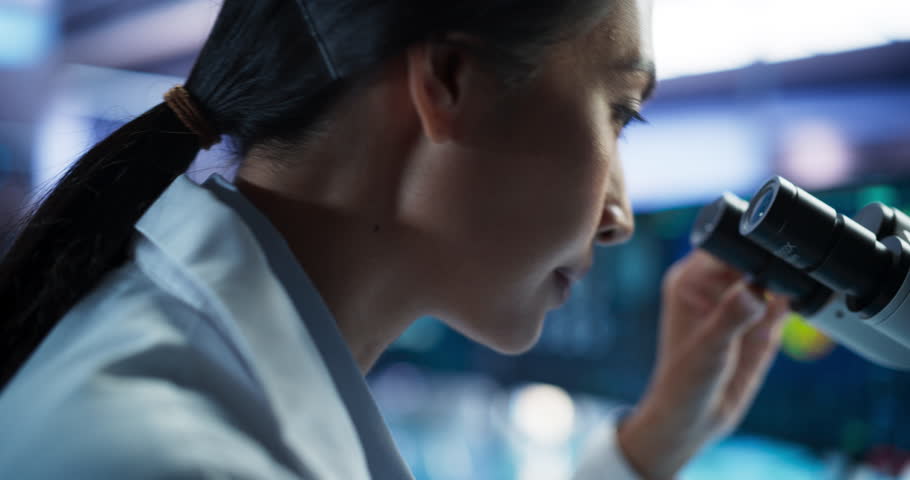 Medical Development Laboratory: Close-Up Portrait of Asian Female Scientist Using Microscope, Analyzes Petri Dish Sample. Big Pharmaceutical Lab doing Medicine, Biotechnology, And Drugs Research.