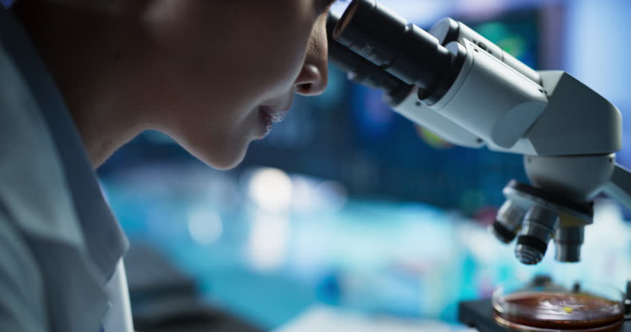 Medical Development Laboratory: Close-Up Portrait of Asian Female Scientist Using Microscope, Analyzes Petri Dish Sample. Big Pharmaceutical Lab doing Medicine, Biotechnology, And Drugs Research.