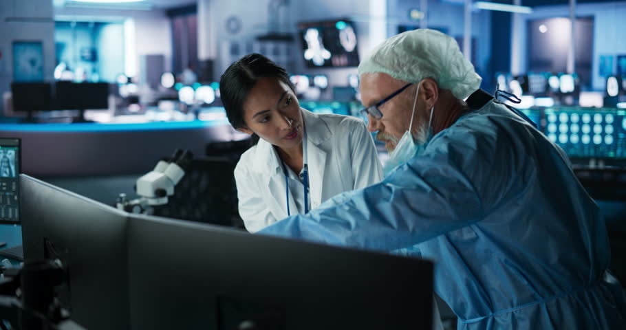 Medical Hospital Research Lab Meeting: Female Asian Neuroscientist Talking To Male Caucasian Neurosurgeon About CT Brain Scans On Desktop Computer. Man Wearing Surgical Gown, Preparing For Surgery. Royalty-Free Stock Footage #1110879725