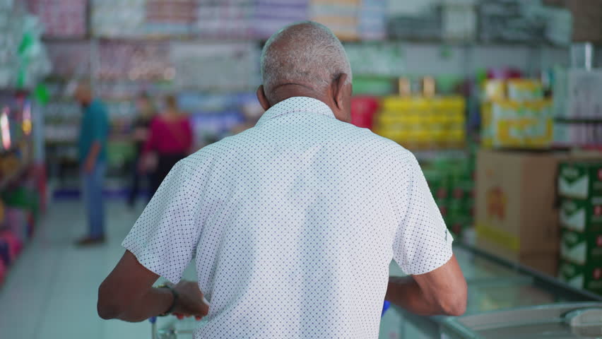 Back of senior black shopper browsing products while walking through grocery store pushing shopping cart