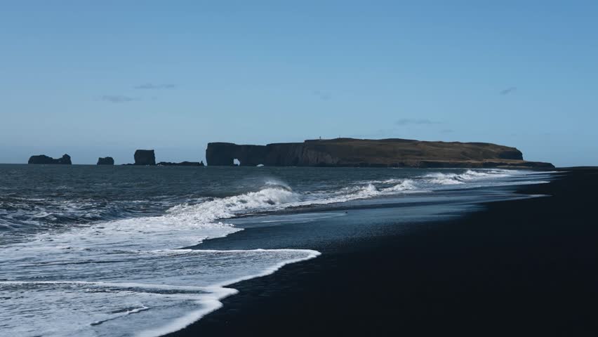 Beauty of Iceland's Black Sand Beach. Rugged landscapes and volcanic serenity. Dyrhólaey promontory in the distance. White waves roll out over the black volcanic sand. Reynisfjara beach. Royalty-Free Stock Footage #1110883953