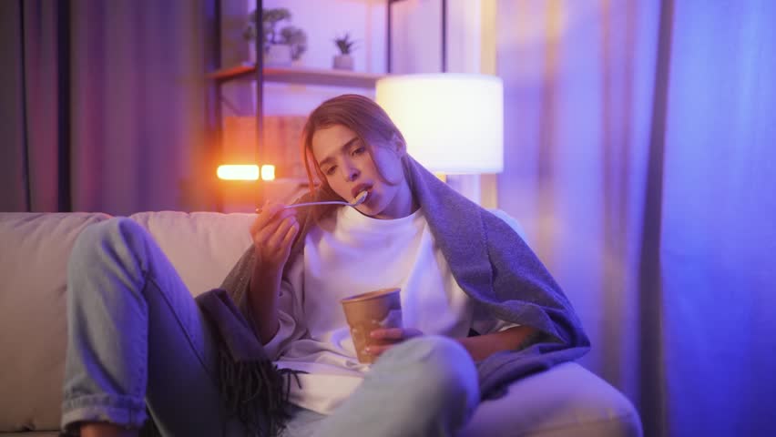 Portrait of sad young woman dealing with stress by eating food sitting on couch at home Upset brunette female wrapped in blanket crying and eating ice cream indoors at late night Mental heath problem Royalty-Free Stock Footage #1110884545