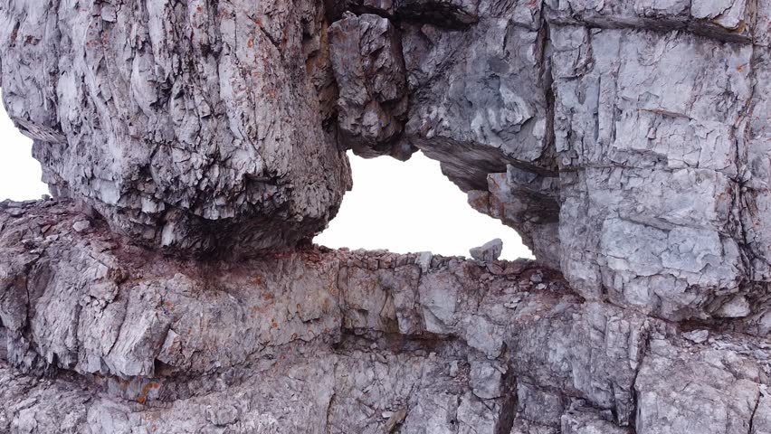 Epic drone aerial flying back from hole in steep rocky stone formation on mountain top in alpine mountains. Foggy mist with summits in distance