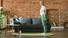 Senior mature woman doing fit exercises with dumbbells while standing on the floor on a mat at home and watching online classes on laptop, workout in living room. Active lifestyle concept