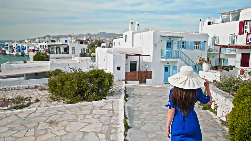 An elegant tourist woman in a blue summer dress with a hat walks through the town of Mykonos Island. Wind blowing the hat of a tourist girl who visits Mykonos, Greece.